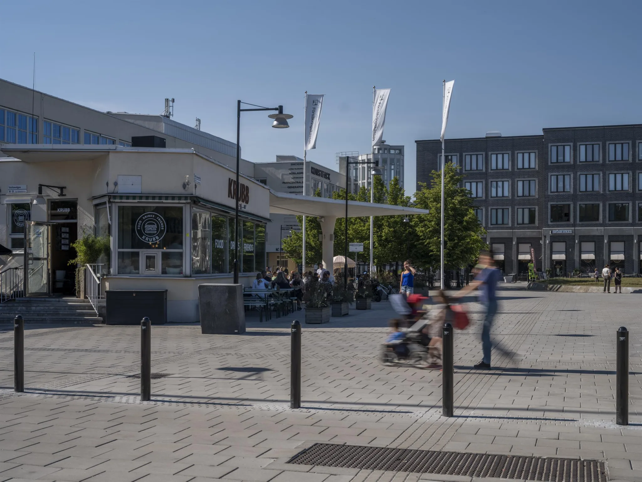 En stadsmiljö med människor som går, en uteservering, byggnader och flaggor i bakgrunden.