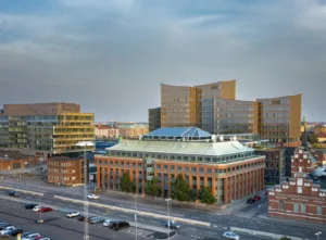 5. Vy över stadsmiljö med en större tegelbyggnad med glasdetaljer och moderna kontorsbyggnader i bakgrunden.