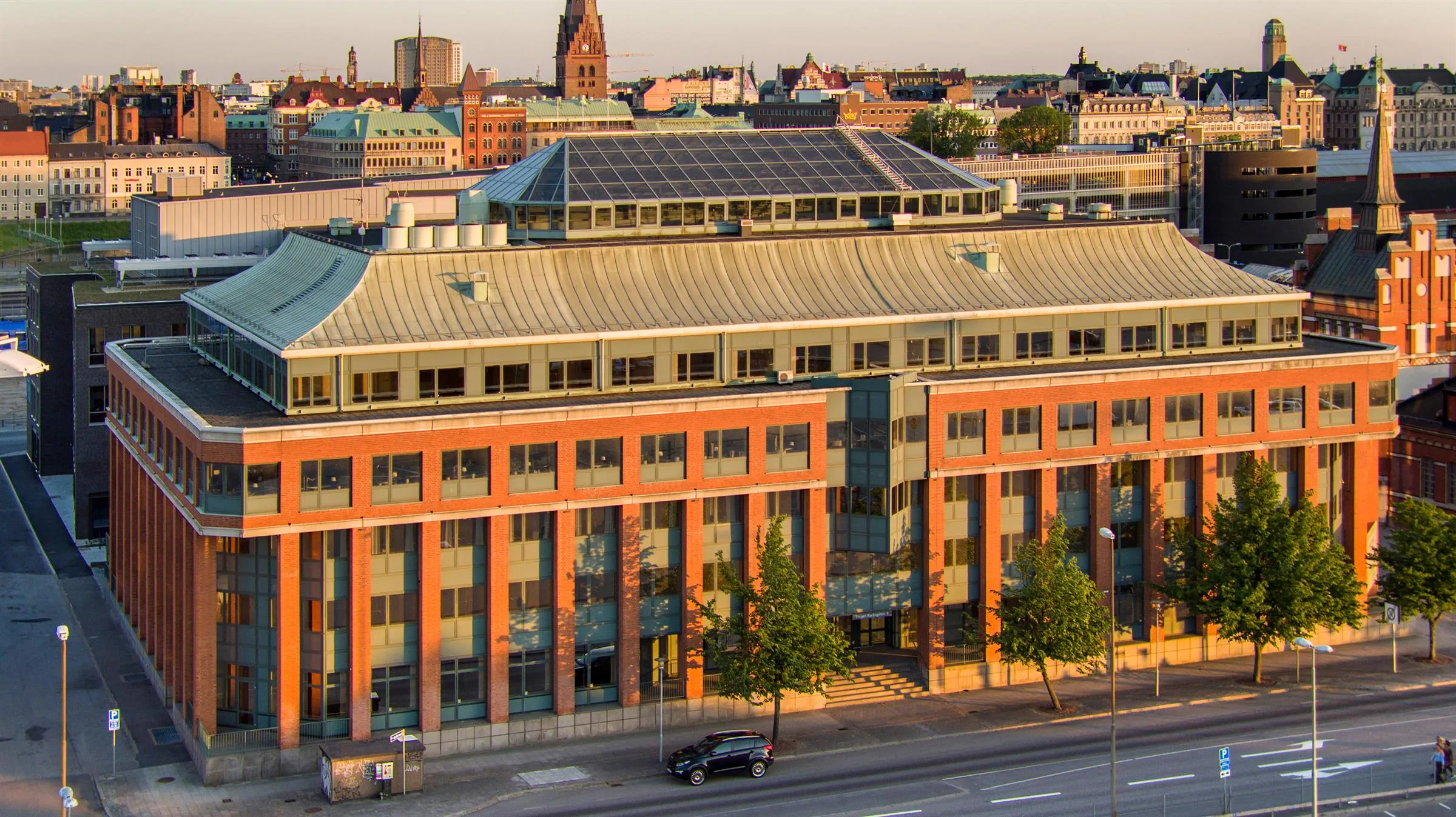 3. Större byggnad i tegel och glas med solpaneler på taket, omgiven av stadsvy i kvällsljus.