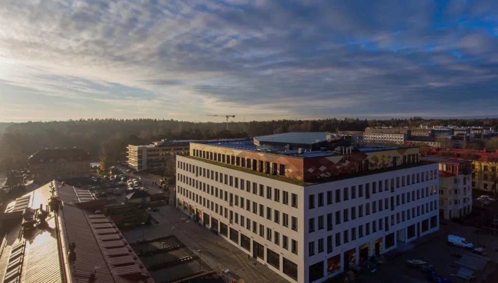 En rektangulär byggnad med vit fasad och stora fönster, omgiven av grönska under en himmel med spridda moln.