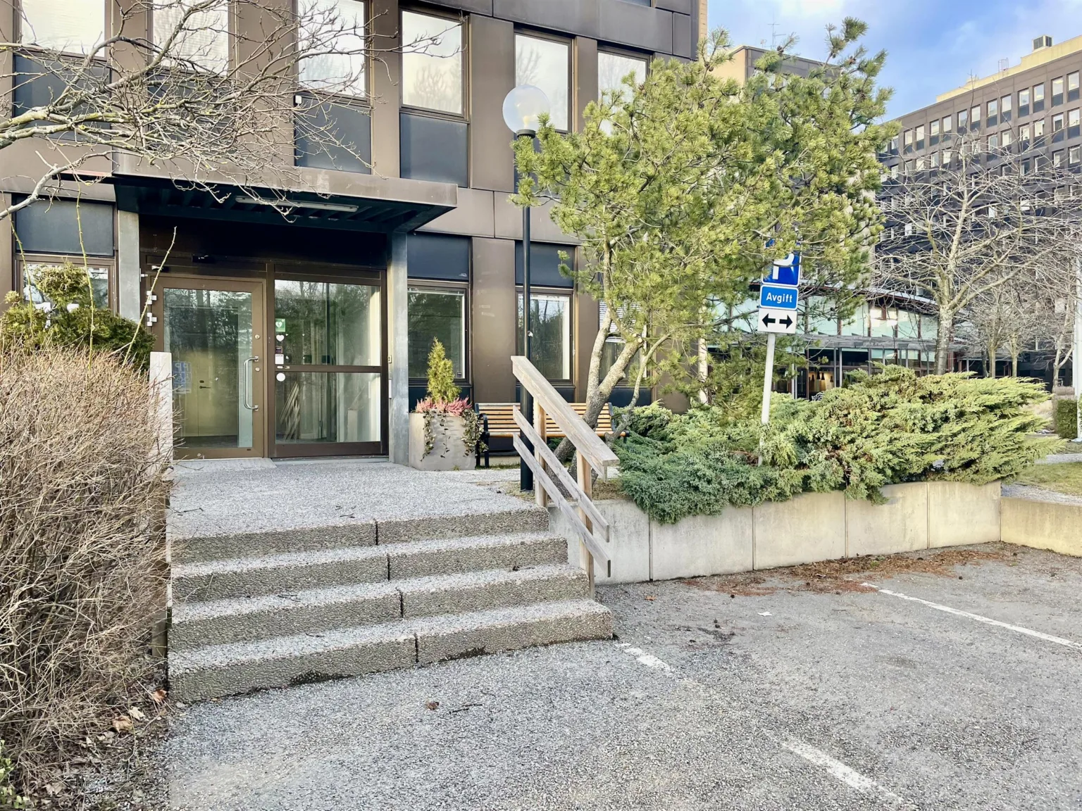 Entré till en byggnad med trappsteg, växtlighet och parkeringsplats.