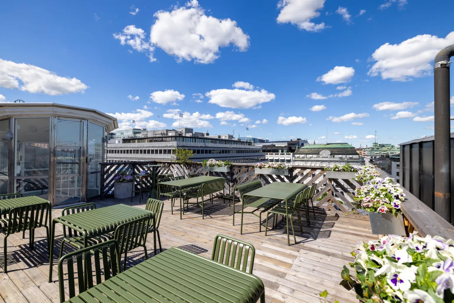 Takterrass med gröna bord och stolar, omgiven av blommor och med utsikt över stadens byggnader under en klarblå himmel med vita moln.