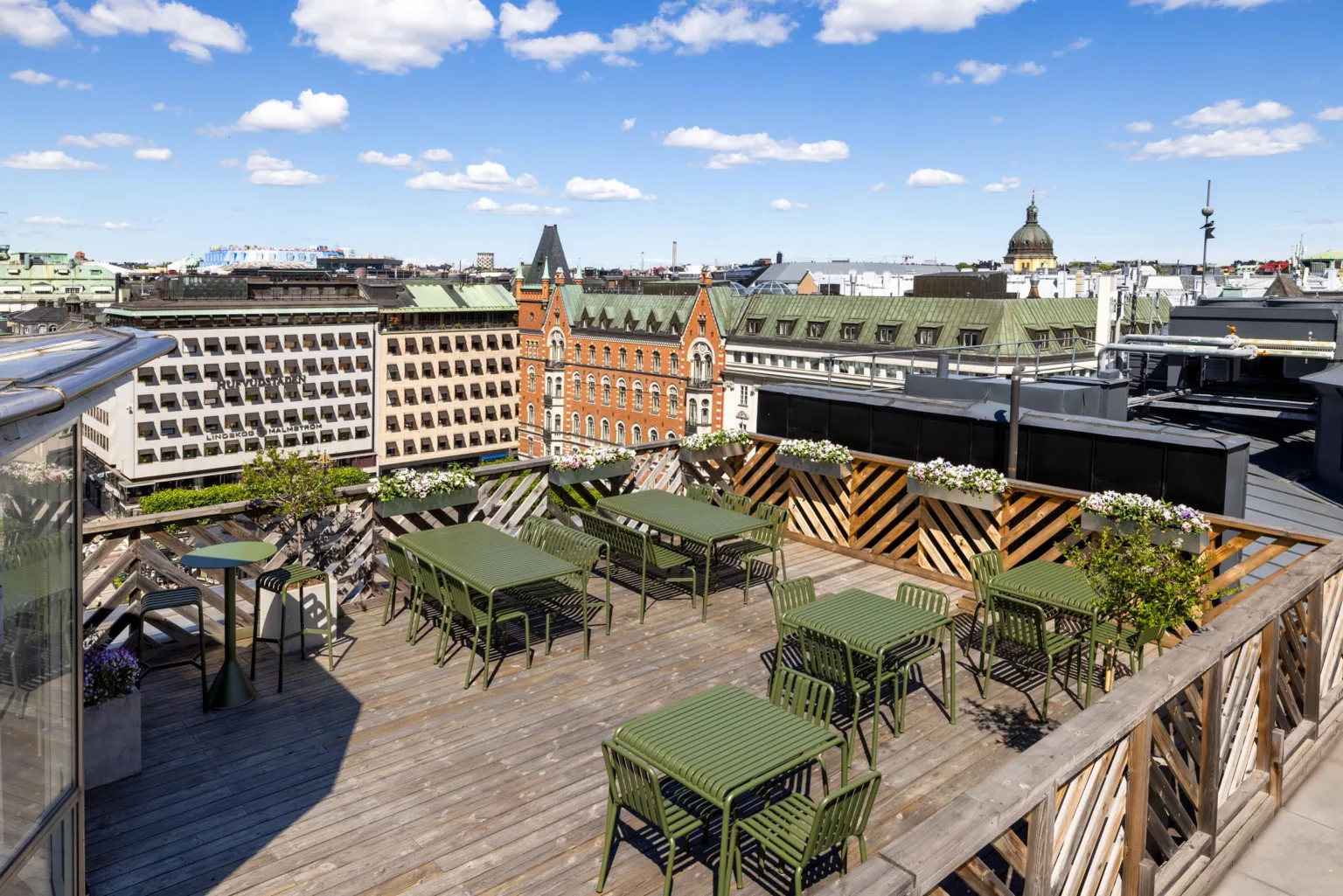 Takterrass med gröna bord och stolar, träplattform och utsikt över stadens äldre och moderna byggnader under en solig dag.