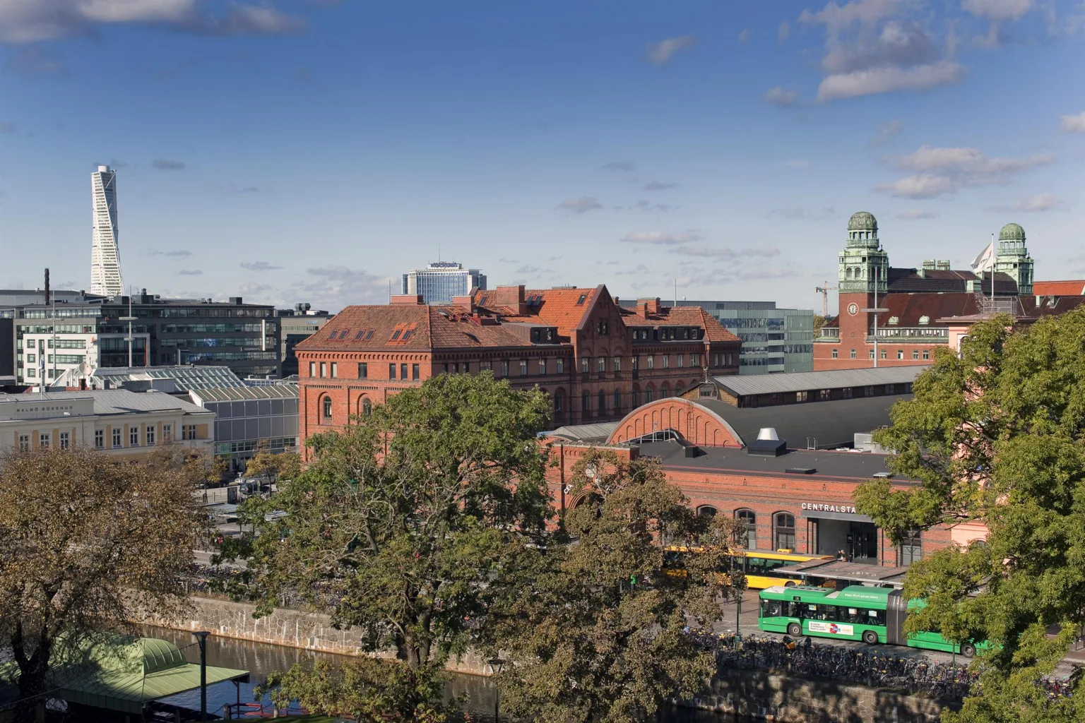 En vy över en byggnad i röd tegel omgiven av gröna träd med moderna kontorsbyggnader i bakgrunden.