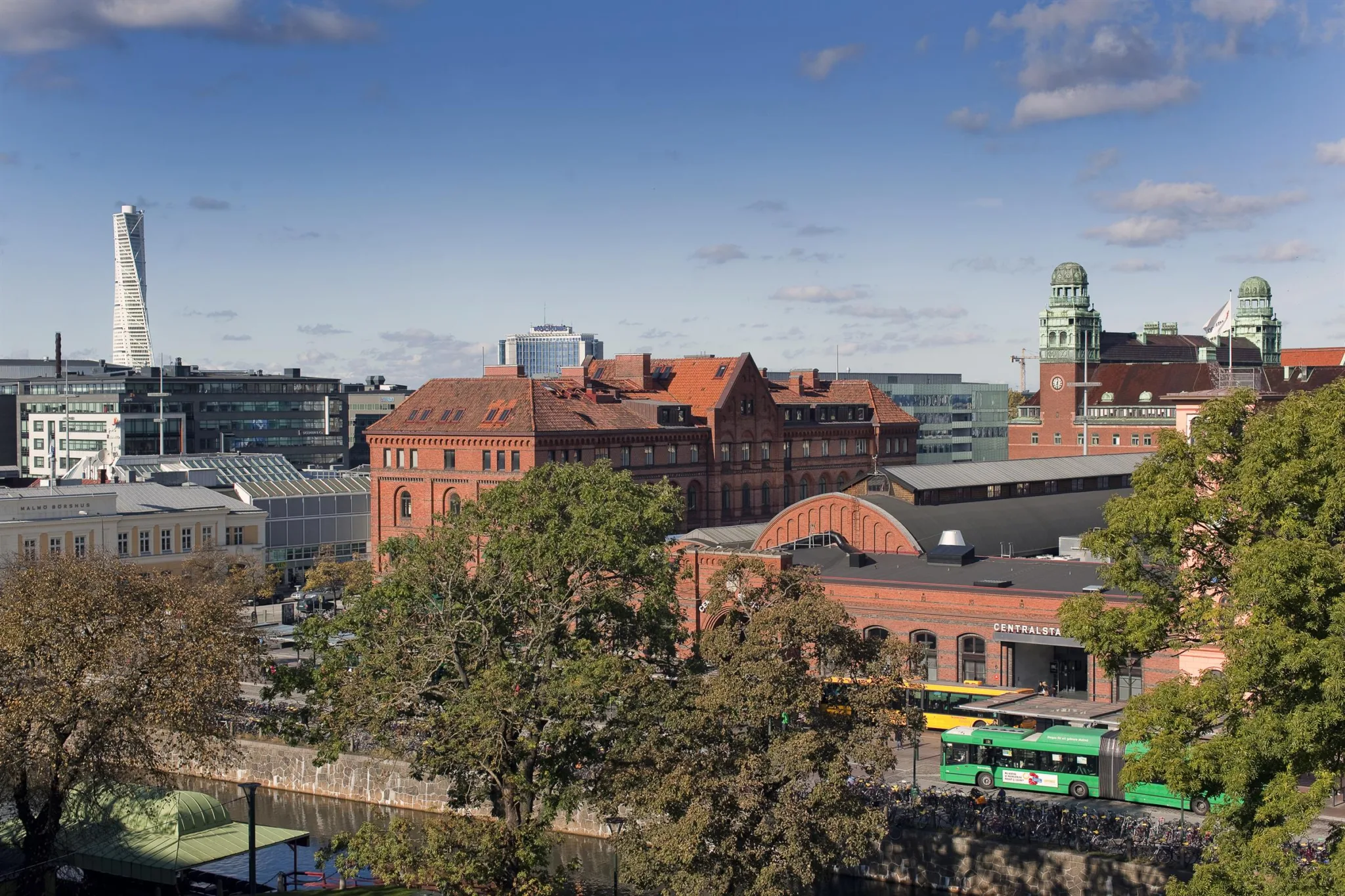 En vy över en byggnad i röd tegel omgiven av gröna träd med moderna kontorsbyggnader i bakgrunden.