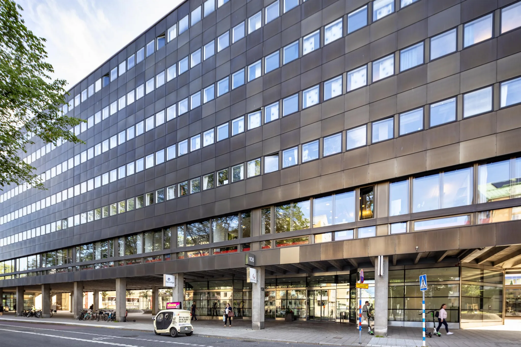 En stor byggnad med flera våningar och glasfönster, med trottoar och bil utanför.