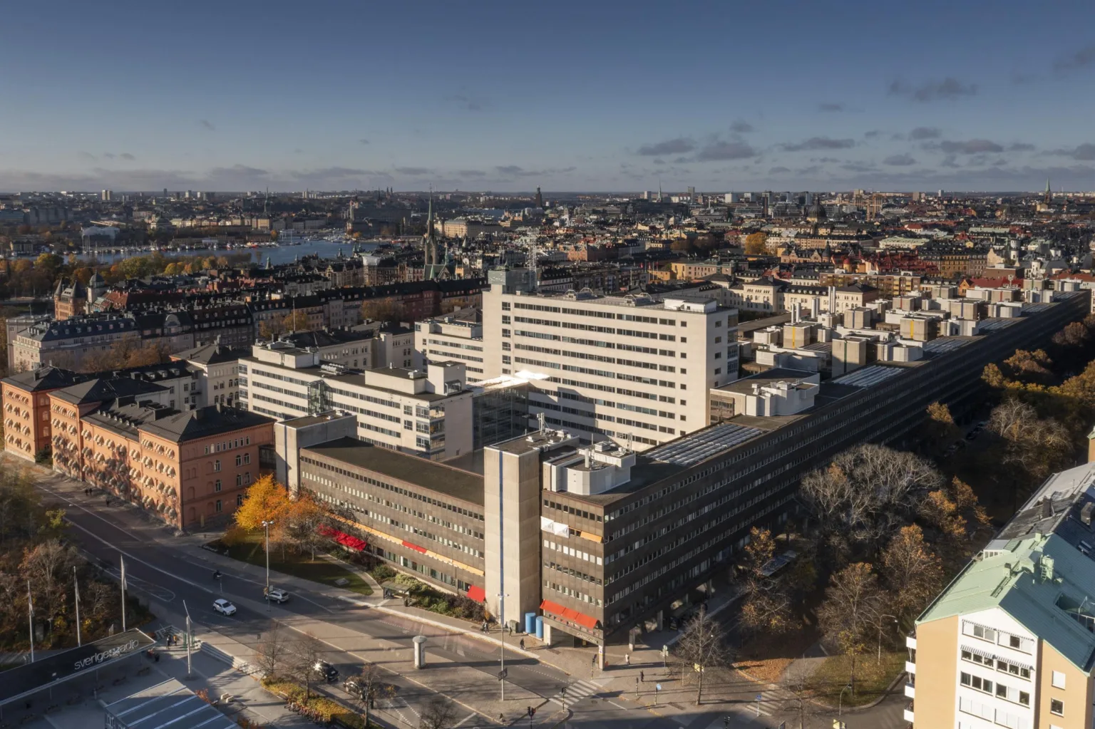 Utsikt över ett stort byggnadskomplex med flera våningar, omgiven av stadsmiljö och vatten i bakgrunden.
