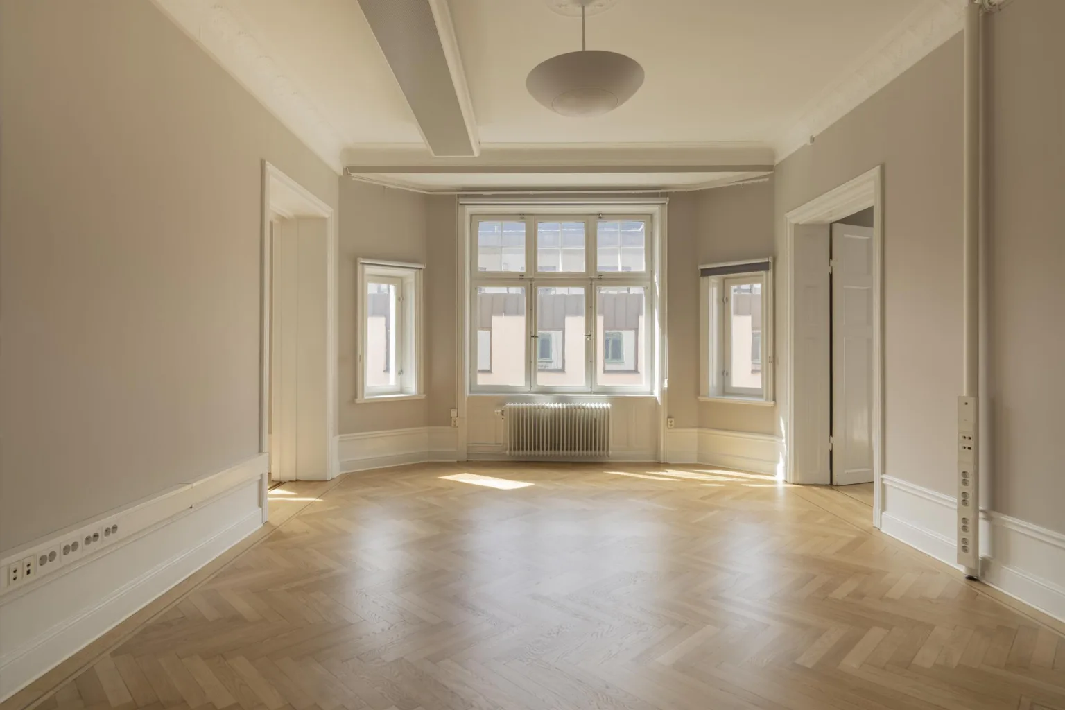 4. Ljust rum med parkettgolv och flera fönster, inklusive ett större fönster med radiator under.