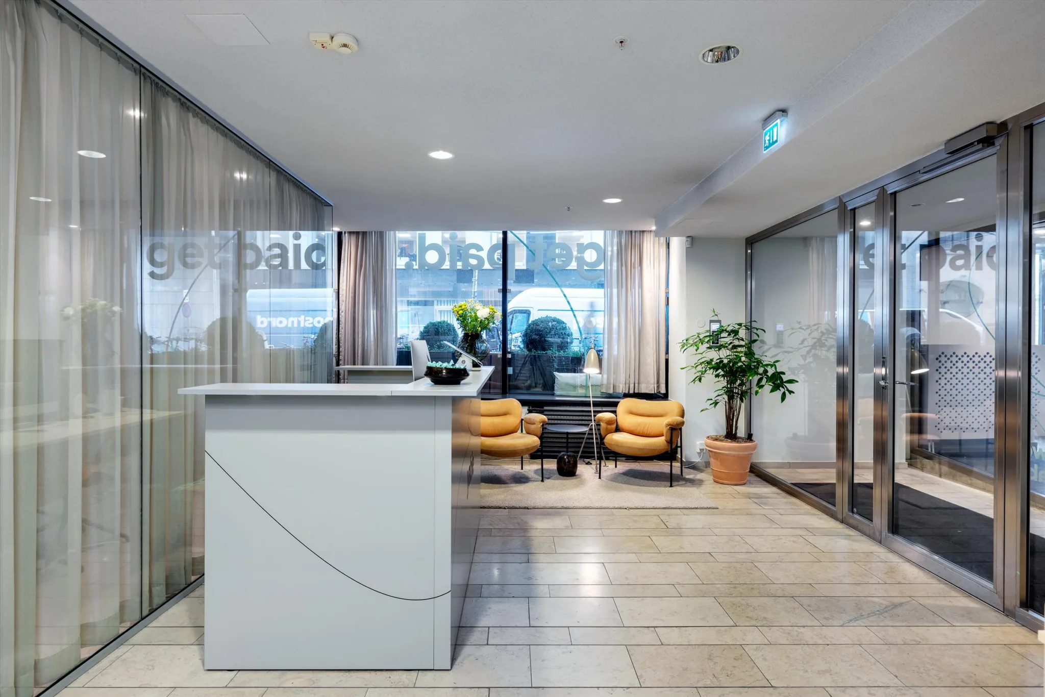 En receptionsyta med en vit disk, två gula fåtöljer och växter vid fönster.