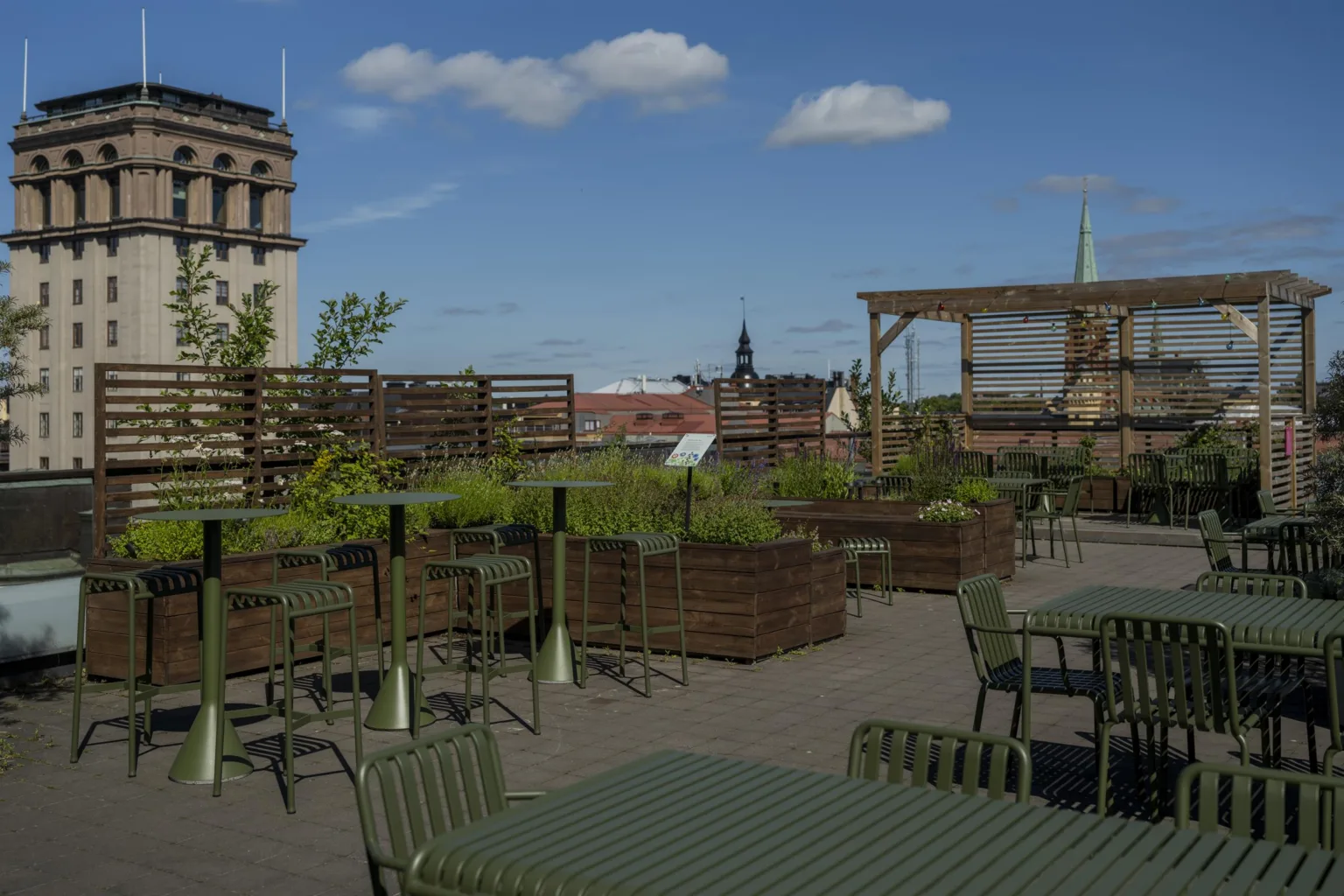 En takterrass med bord, stolar, grönska och utsikt över stadens byggnader under en blå himmel.