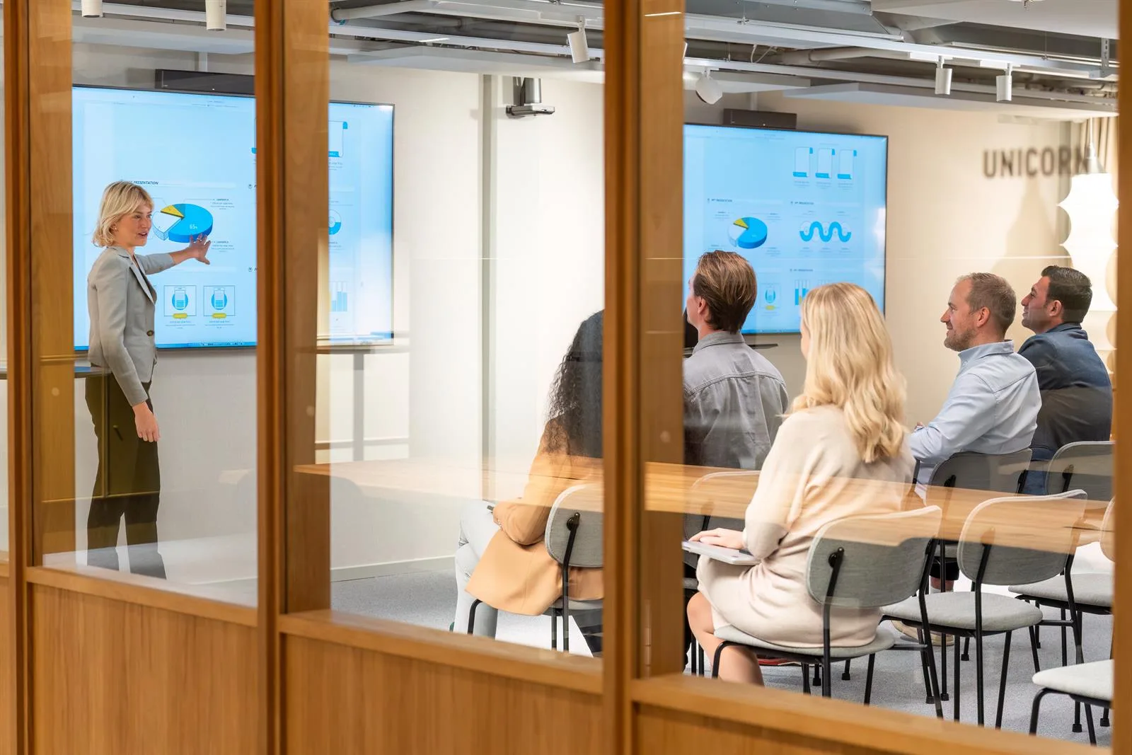 En grupp människor sitter vid ett konferensbord och tittar på presentationer på två skärmar, sett genom glasväggar.