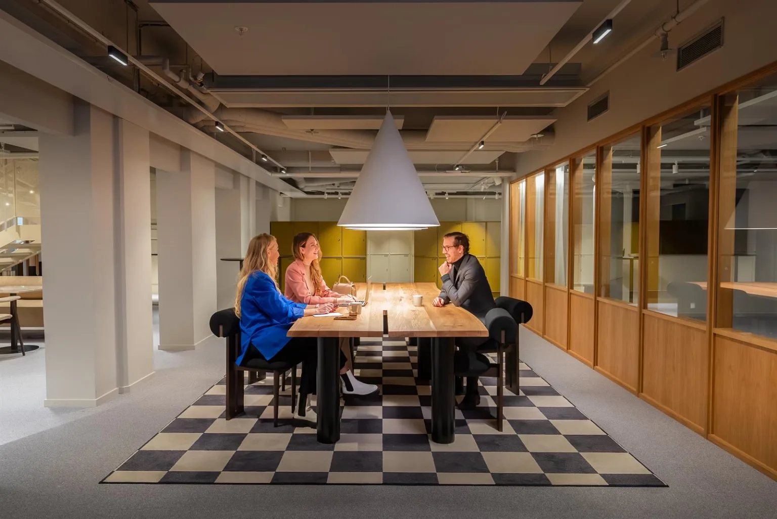 Tre personer sitter vid ett stort bord i ett kontorsutrymme med modern inredning och en stor lampa ovanför.