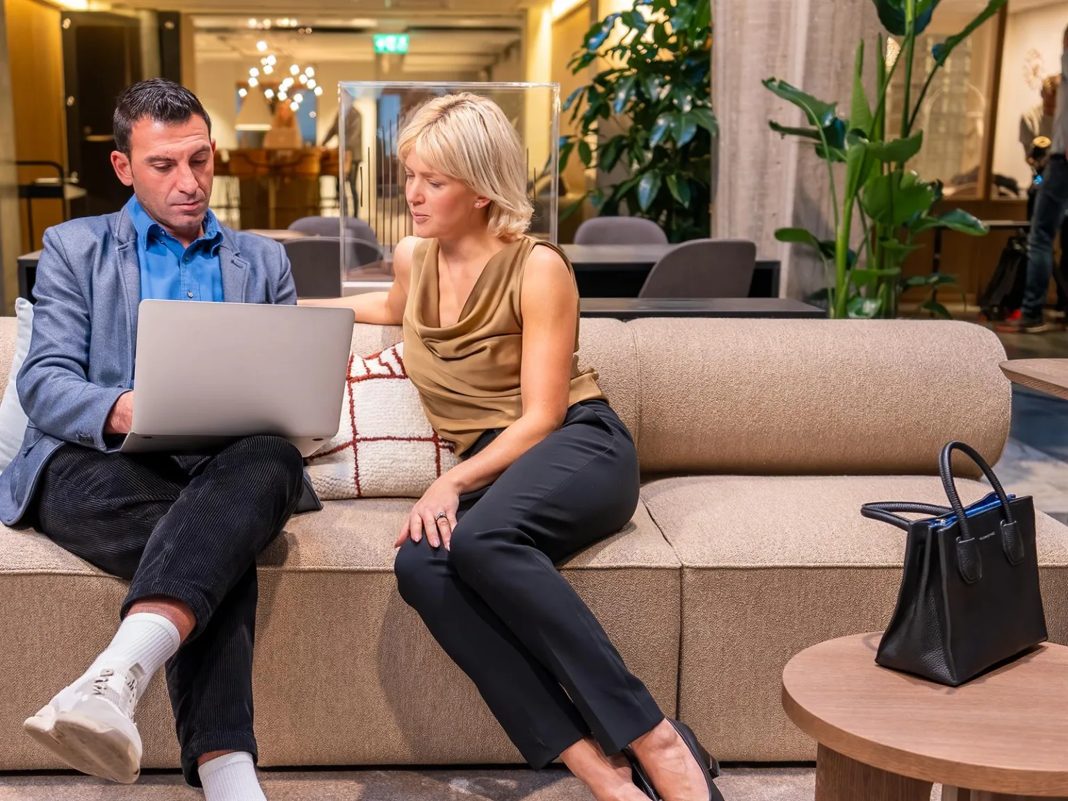 Två personer sitter på en beige soffa och tittar på en bärbar dator, med en växtfylld bakgrund.