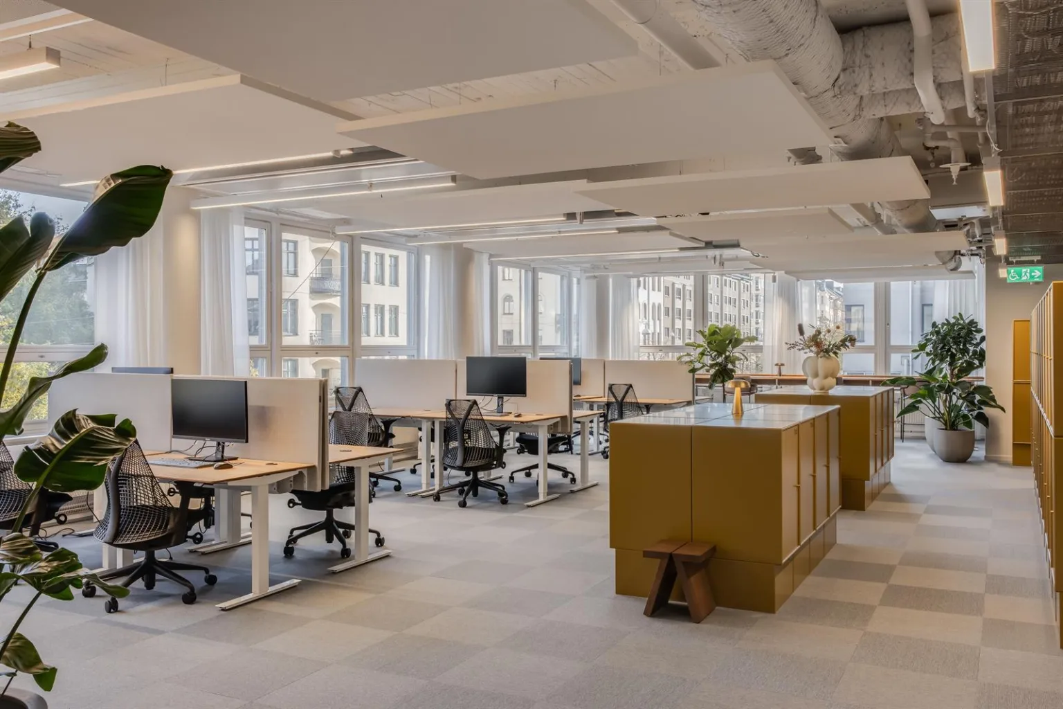 Ett öppet kontorslandskap med flera arbetsstationer, stora fönster och en central köksö med dekorationer.