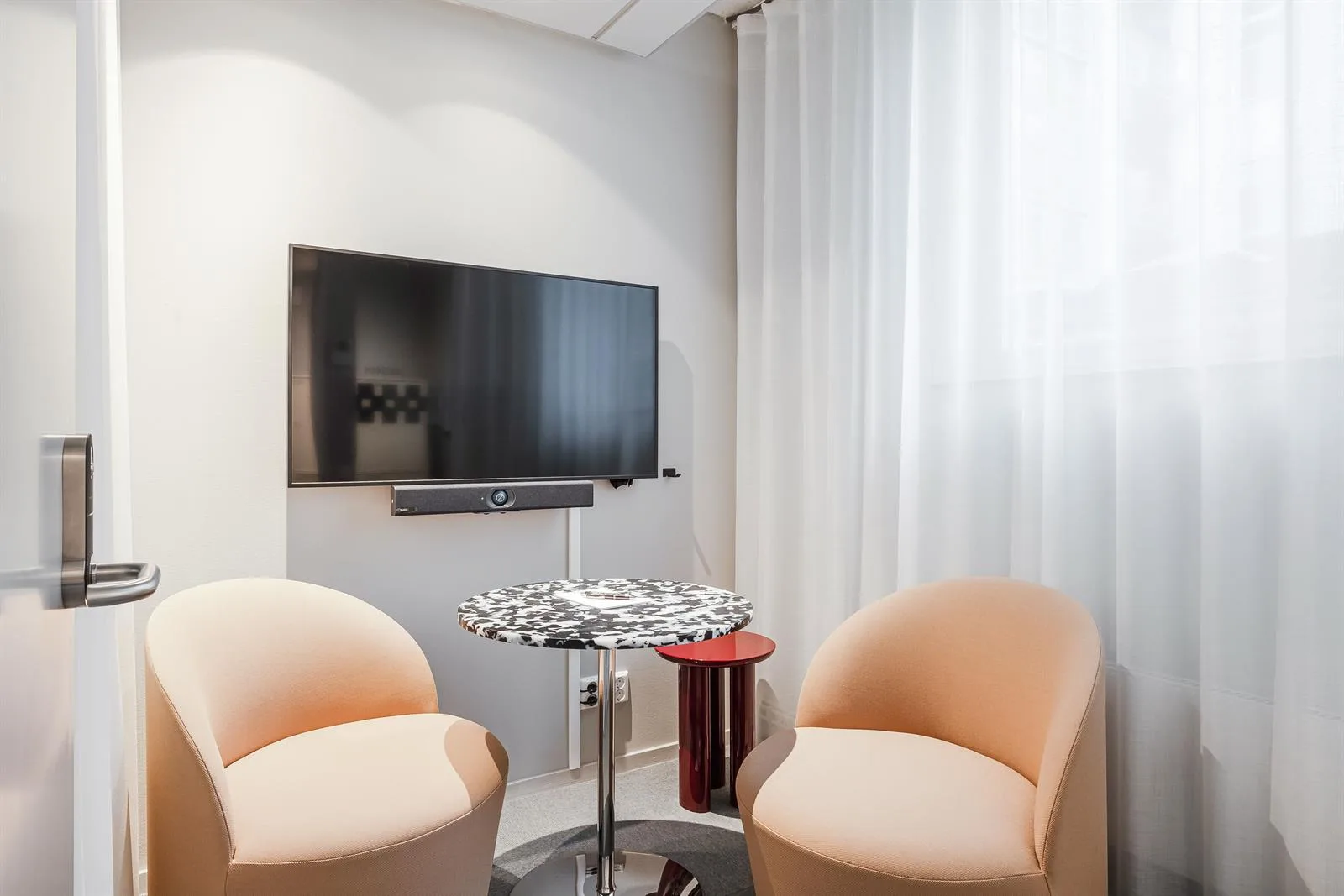 Två beige fåtöljer står runt ett litet bord framför en väggmonterad TV och vit gardin.