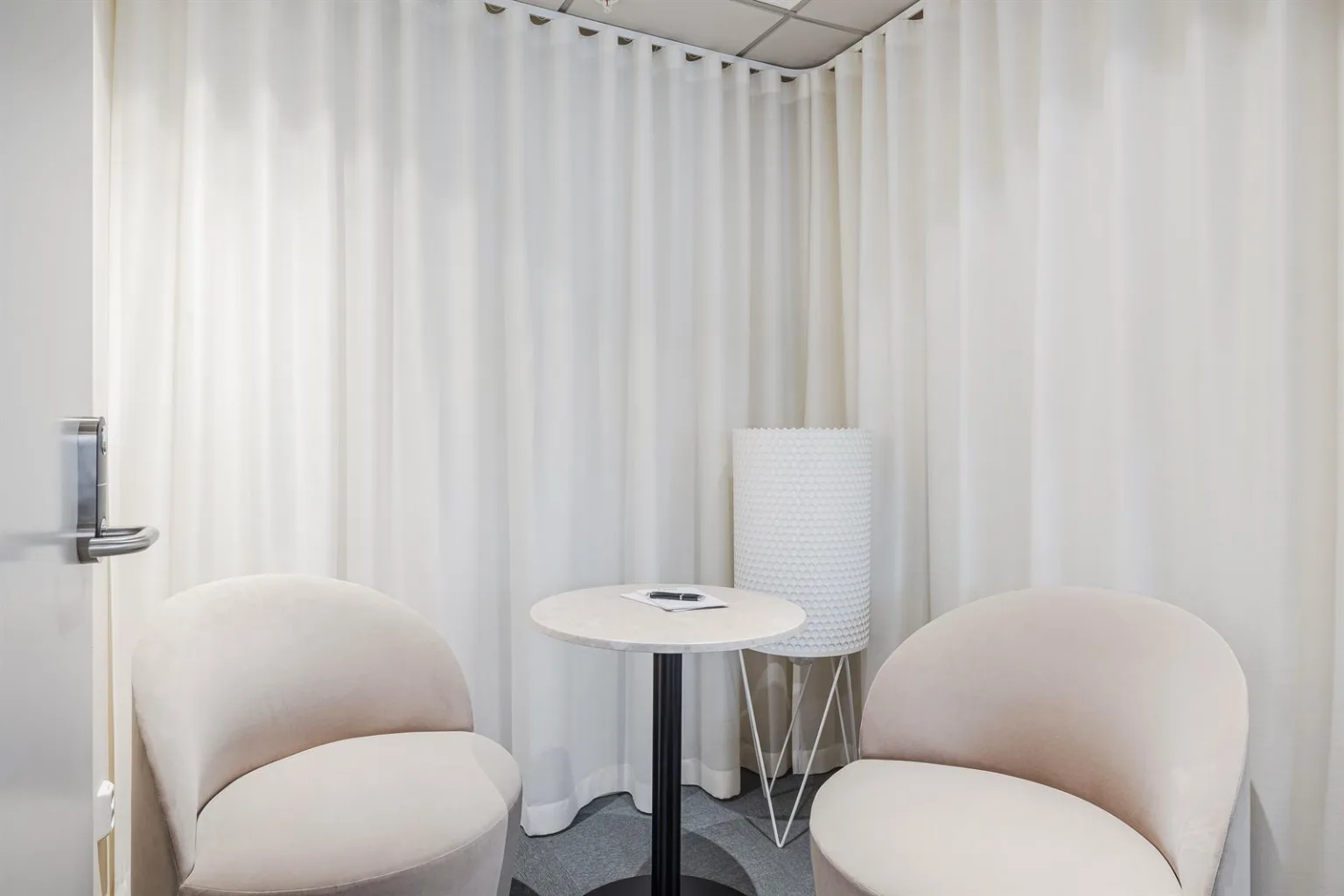 Två fåtöljer och ett litet bord framför vita gardiner, med en modern golvlampa i hörnet.