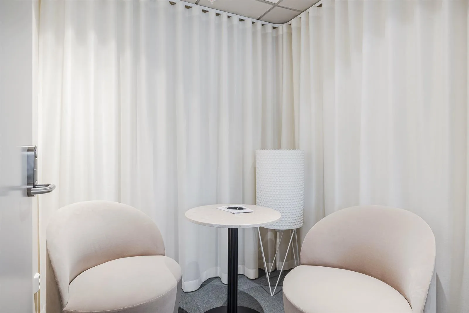 Två fåtöljer och ett litet bord framför vita gardiner, med en modern golvlampa i hörnet.