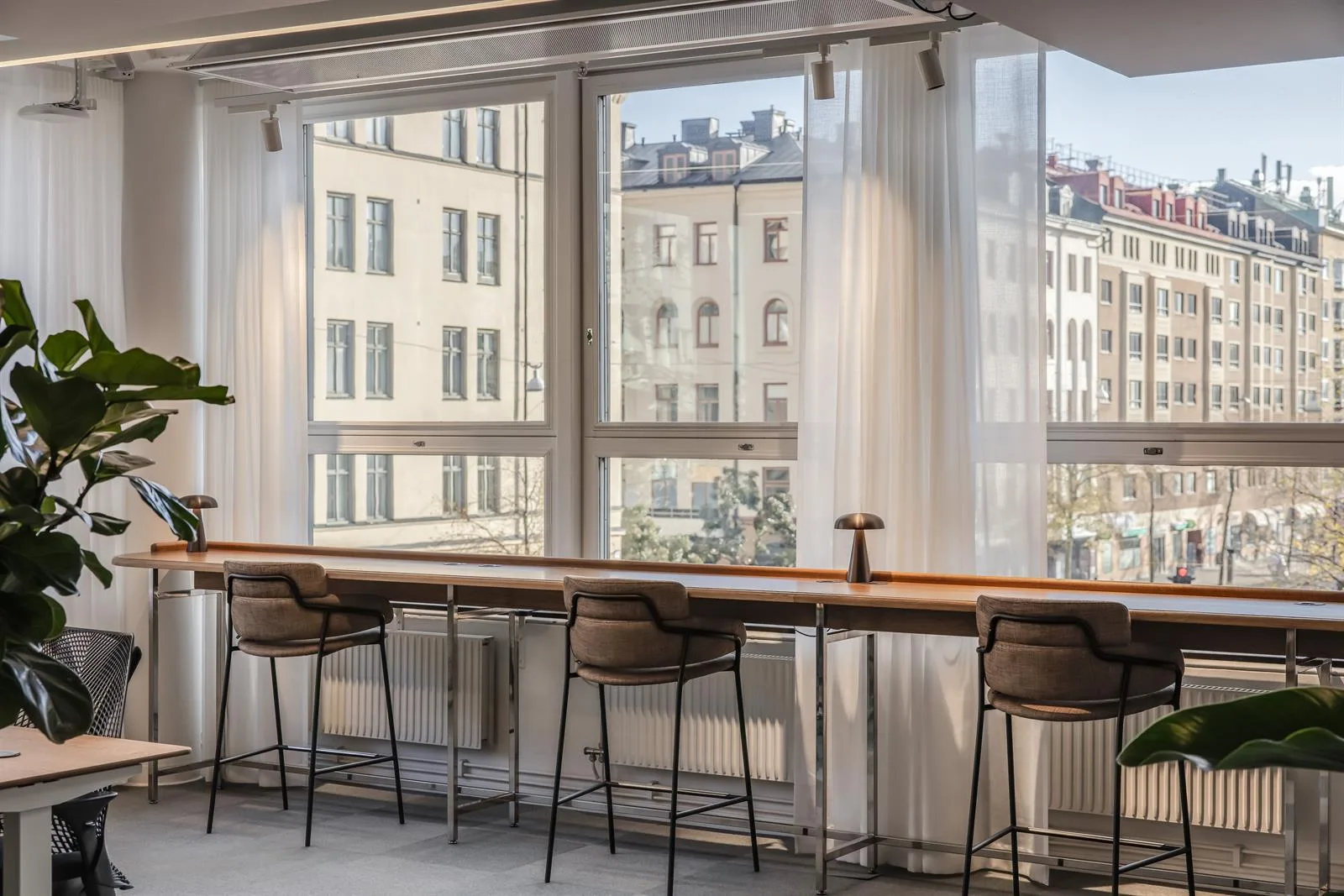 En lång arbetsbänk med höga stolar står vid stora fönster som släpper in dagsljus, med utsikt över byggnader.