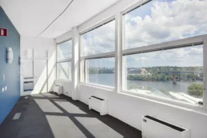 Ett ljust kontorsrum med stora fönster som erbjuder utsikt över vatten och grönska, med solljus som lyser in.