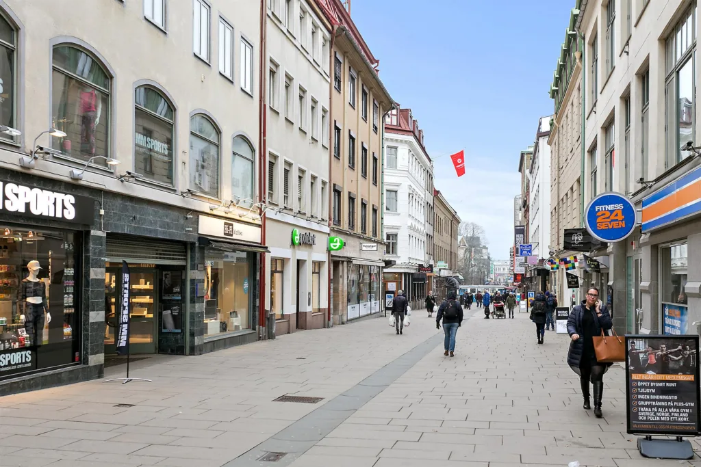 En stadsgata kantad av butiker och byggnader med fotgängare som promenerar på gångvägen under en klar himmel.