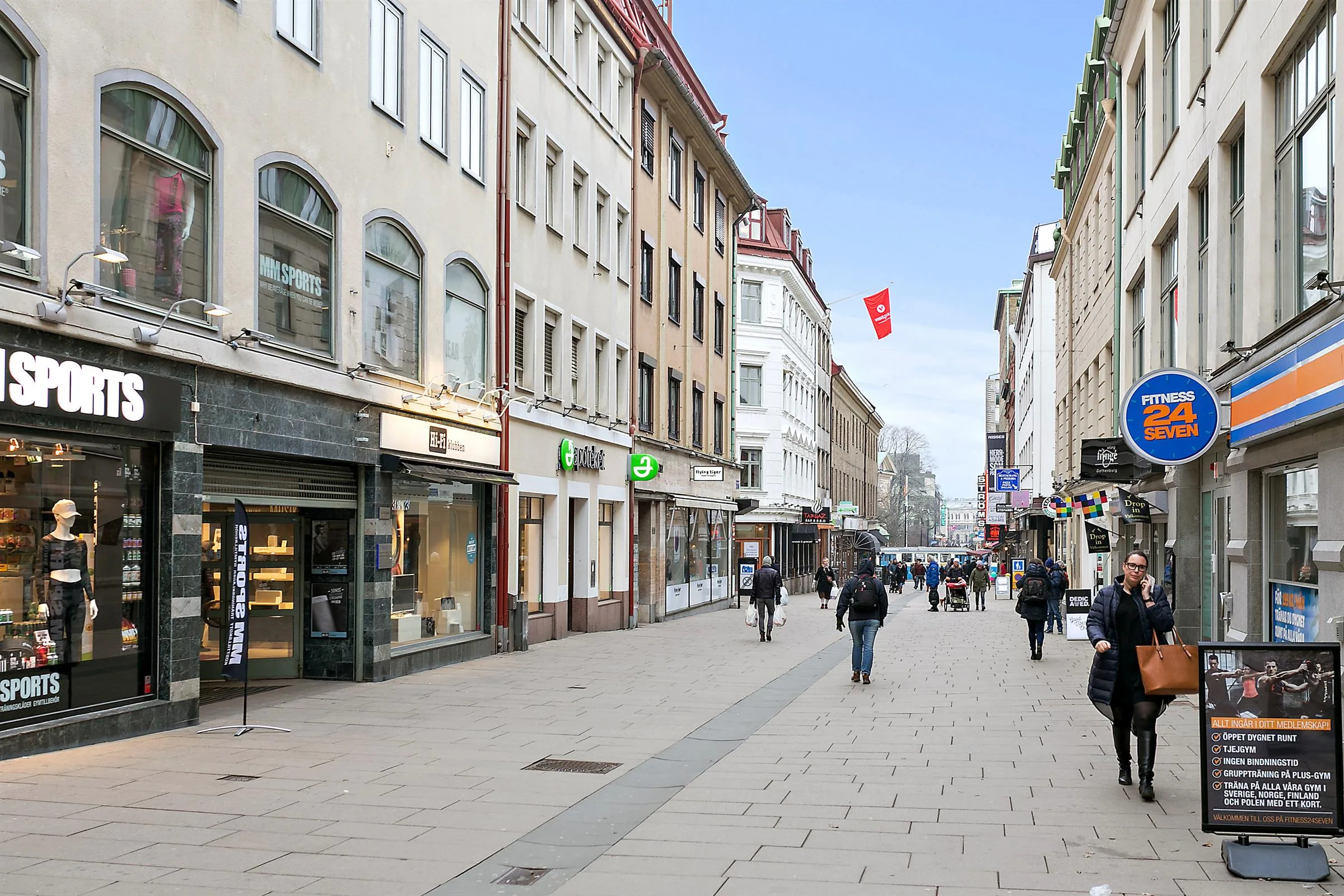En stadsgata kantad av butiker och byggnader med fotgängare som promenerar på gångvägen under en klar himmel.