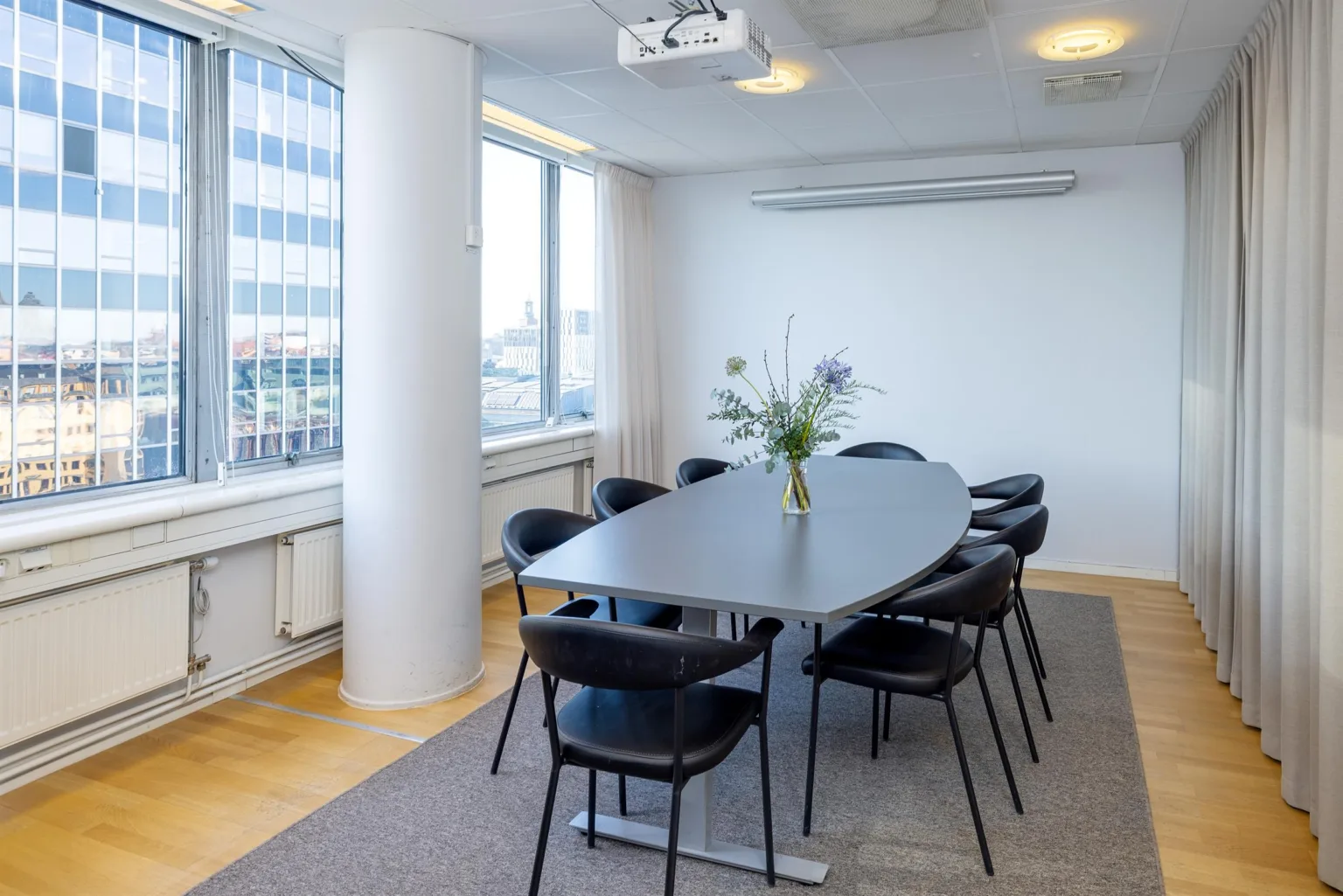 Ett konferensrum med ett ovalt bord, åtta svarta stolar, en vas med blommor och stora fönster.