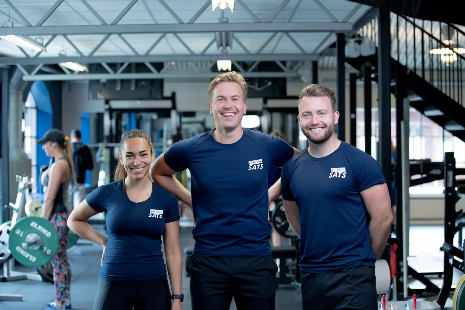 Tre personer i träningskläder står och ler inuti ett gym, med träningsutrustning i bakgrunden.