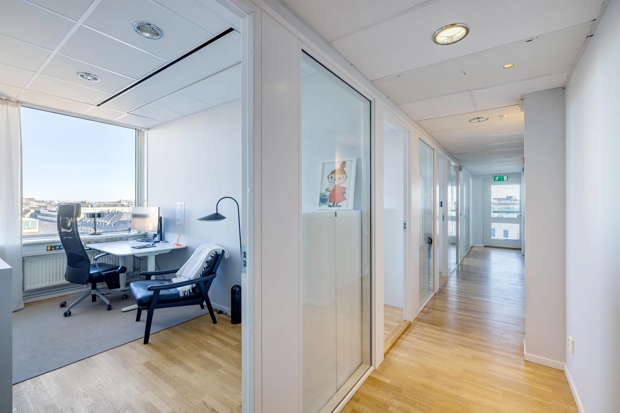 En kontorskorridor med glasväggar, ett arbetsrum med skrivbord och stol till vänster.