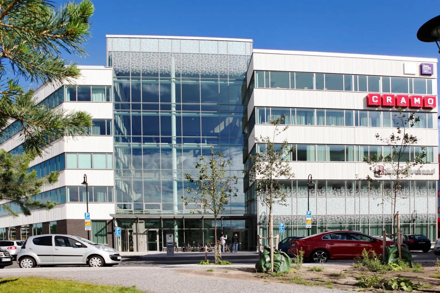 En vit byggnad med glasentré och parkerade bilar framför, omgiven av träd och blå himmel.