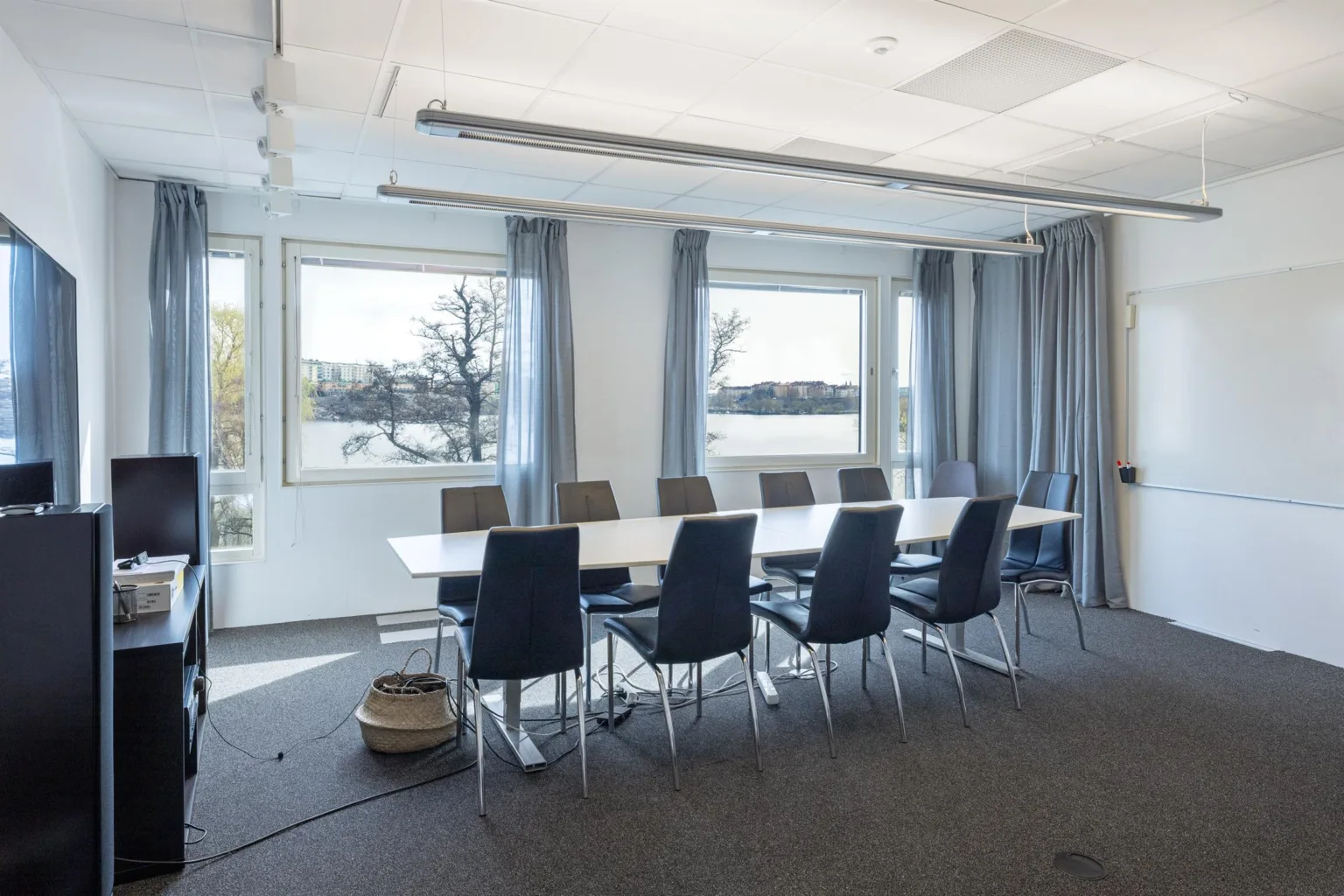 1. Ett konferensrum med ett långt bord, åtta stolar och stora fönster som visar en utsikt över vatten och träd.